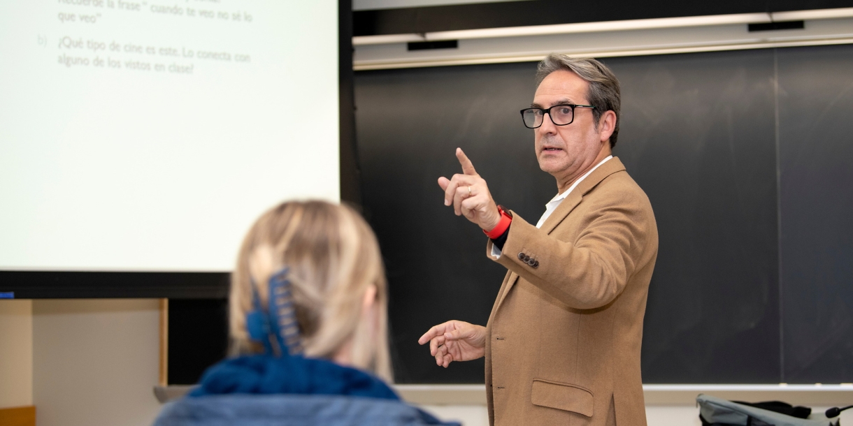 Professor Fernando Blanco stands in front of a chalkboard and projector screen, raising a finger as he makes a point during a class lecture